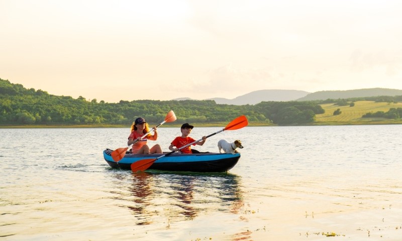 Dete vesla pored žene i psa Džek Rasela u čamcu na naduvavanje preko mirne vode u zalazak Sunca